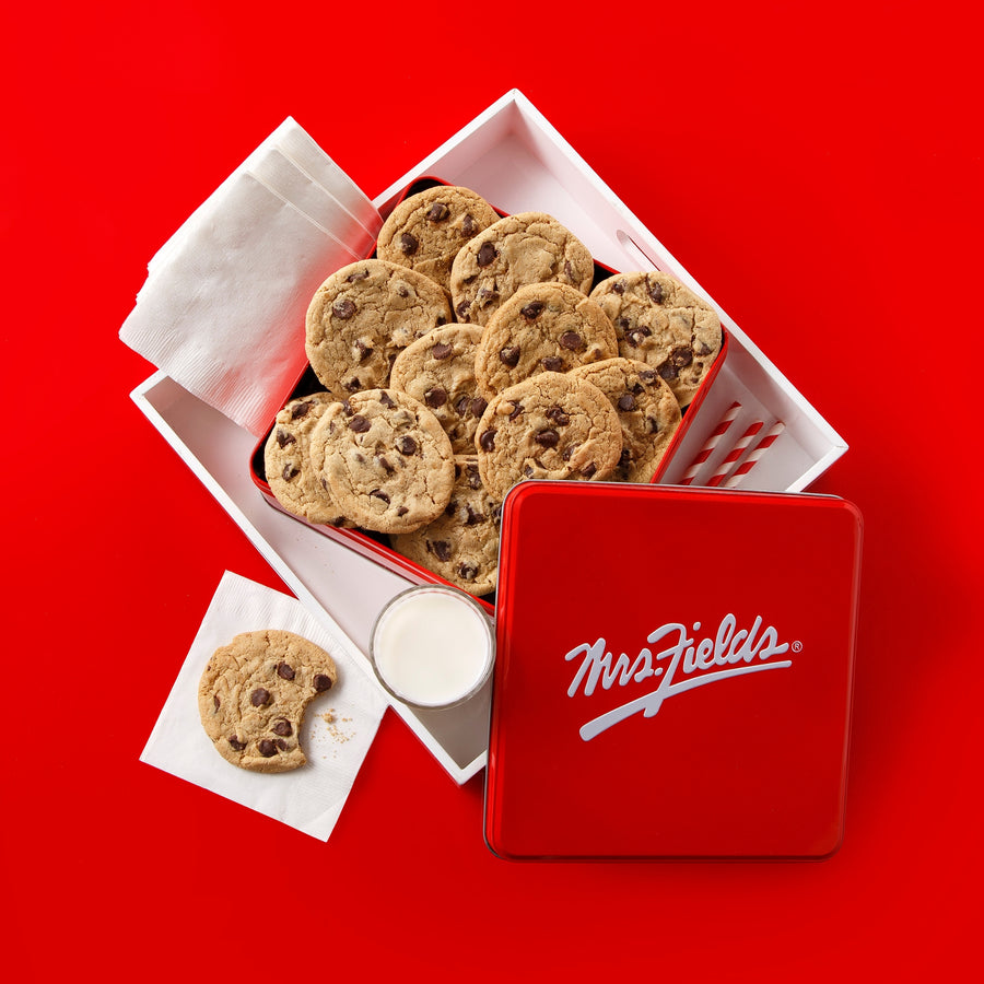 A red tin of Mrs. Fields chocolate chip cookies arranged in an open white box on a bright red background, with a few cookies on napkins and a small cup of milk beside the box.