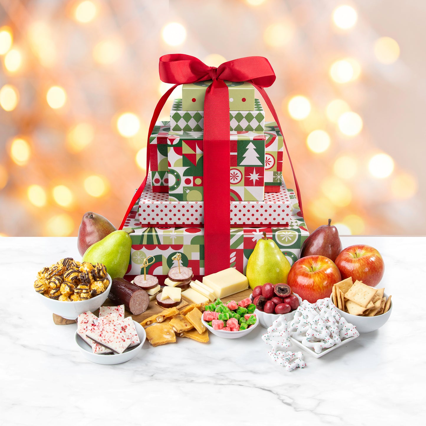 A multi-tiered gift tower surrounded by an assortment of fruit, popcorn, frosted pretzels, peanut brittle, candy, crackers, cheese, sausage, and peppermint bark.