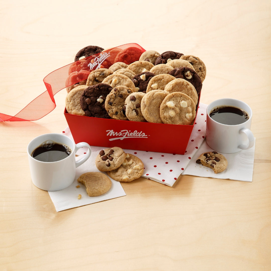 A small red Mrs. Fields crate filled with bite-sized cookies in a variety of flavors on a light wooden surface with a polka dot napkins and two white mugs of coffee.