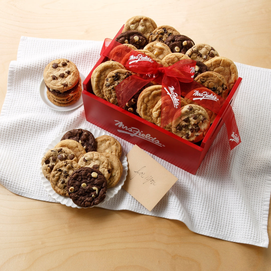 A red wooden gift crate filled with assorted chocolate chip cookies, tied with a red ribbon, displayed on a white textured cloth on a light wood table. Several cookies are arranged on white plates in front, with a small card in front of the box.