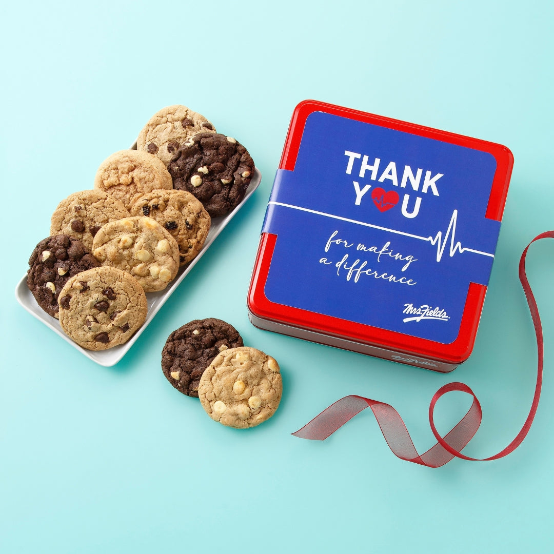 A square red and blue cookie tin with a ribbon band reading 'THANK YOU for making a difference' and the TruChocolate logo, shown next to a white plate of assorted chocolate chip cookies on a light aqua background.