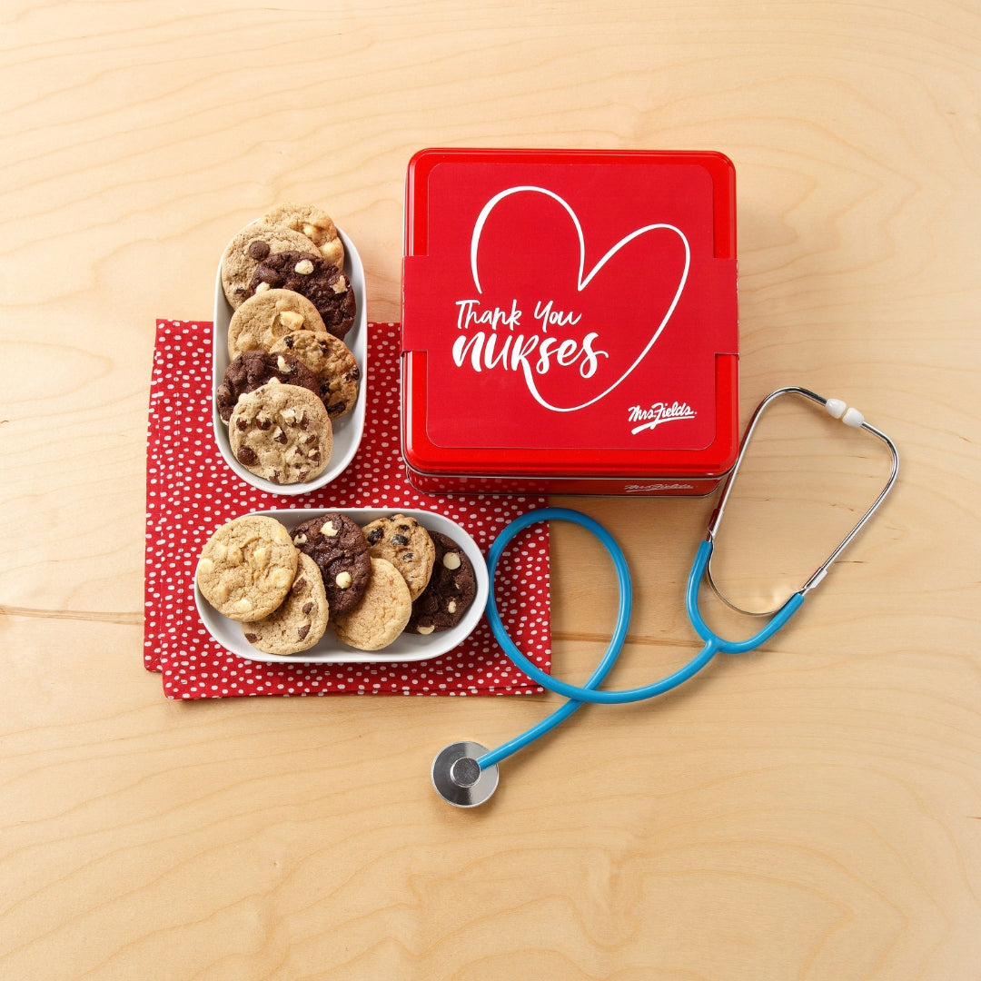 A red square gift tin with a white heart outline and the words 'Thank You Nurses' on the lid, styled on a light wood surface with two white bowls of cookies and a blue stethoscope.