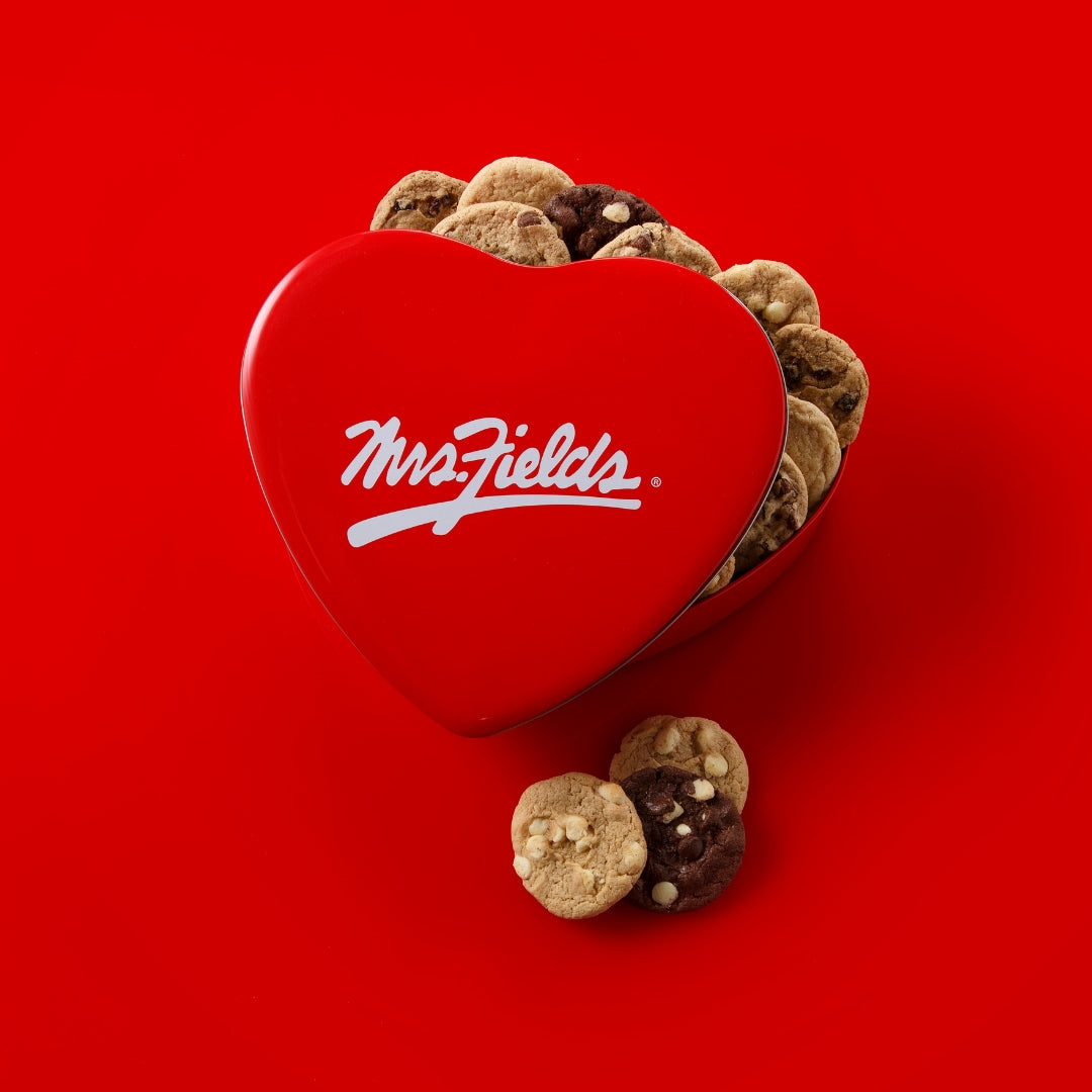 A red heart-shaped metal cookie tin with the white Mrs. Fields logo, filled with assorted cookies on a solid red background.