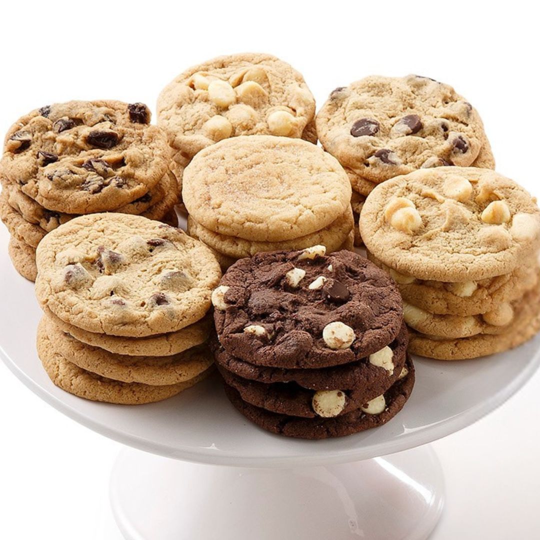 A variety of chocolate chip cookies in different flavors, stacked on a white ceramic pedestal plate against a clean white background.