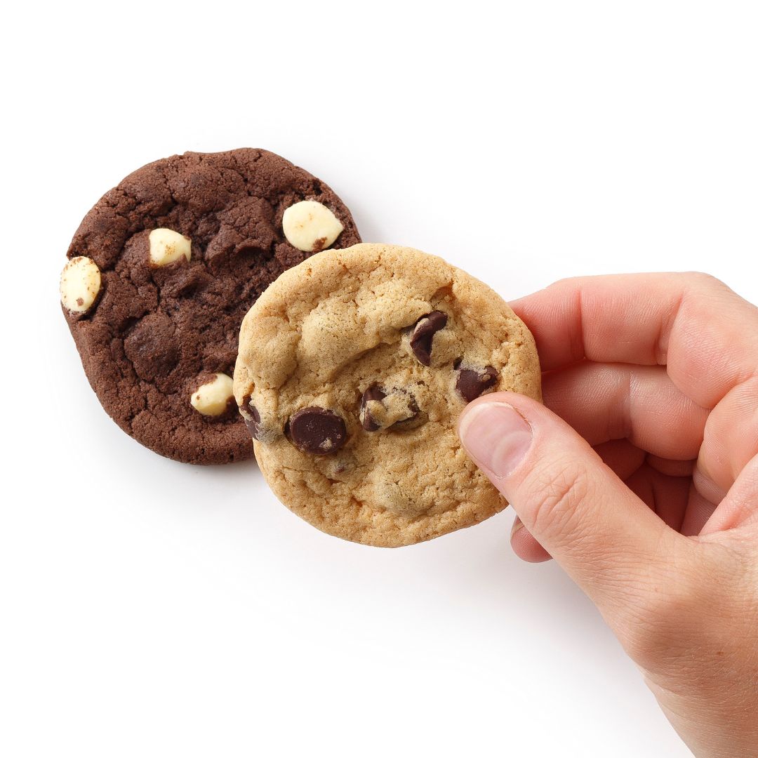 Two cookies on a white background, one dark chocolate cookie and one lighter chocolate chip cookie being held by a person's hand.