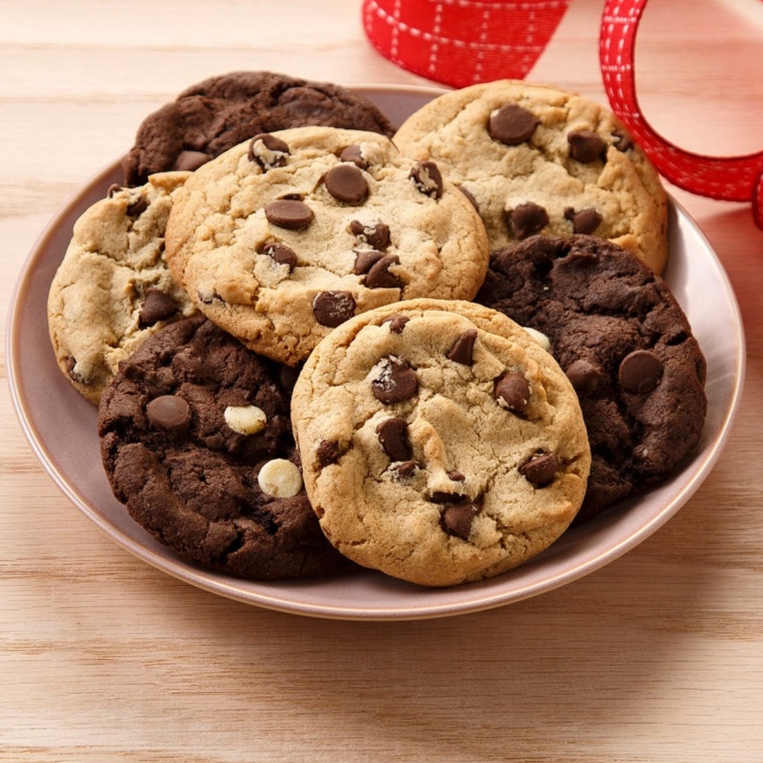 A plate of assorted chocolate chip cookies on a light wood table, with a red patterned ribbon partially visible in the background.