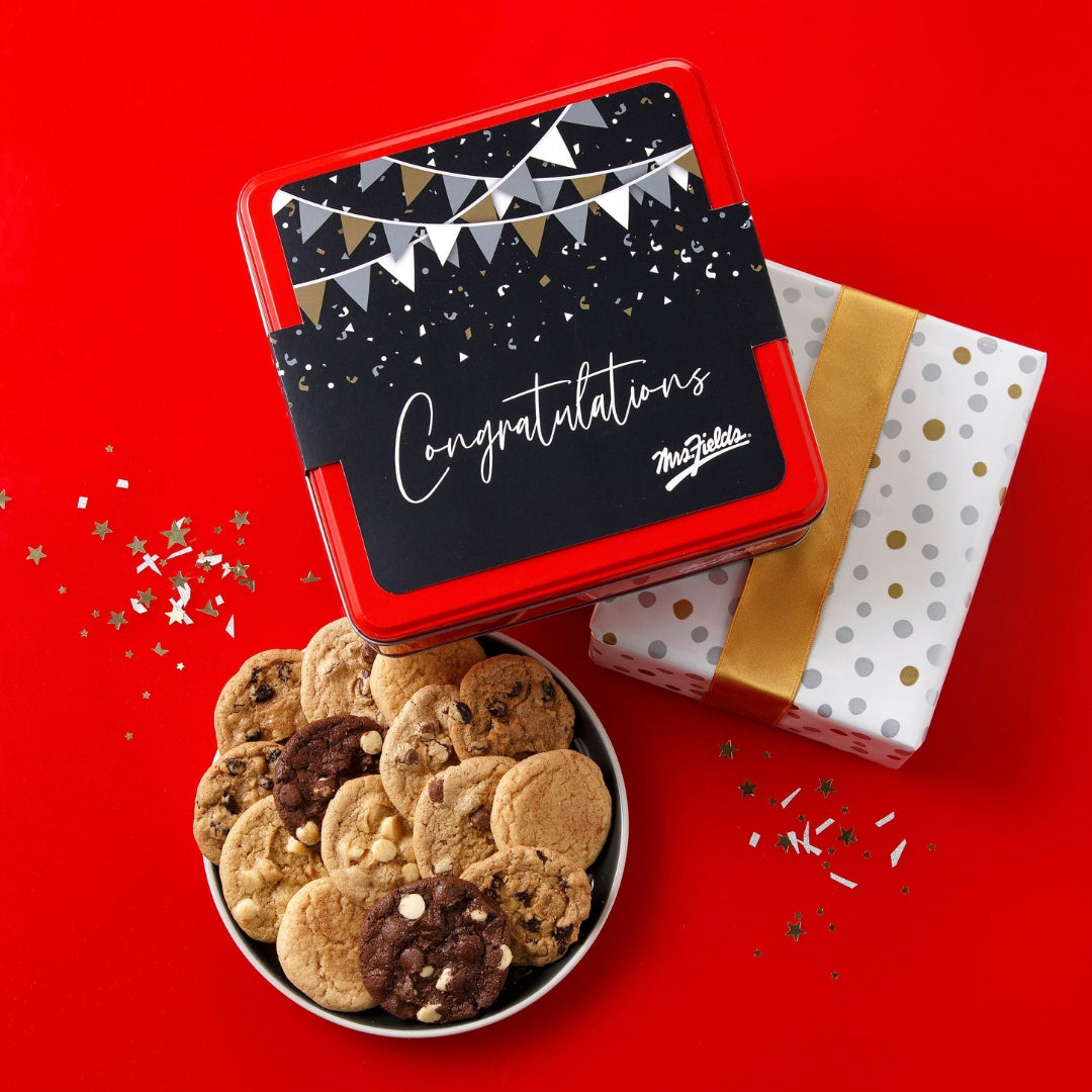 A red metal cookie tin with a black 'Congratulations' label sits on a bright red background, with a bowl of assorted cookies in front and a wrapped gift box behind it. The script logo 'The Cookie Club' is visible on the label.