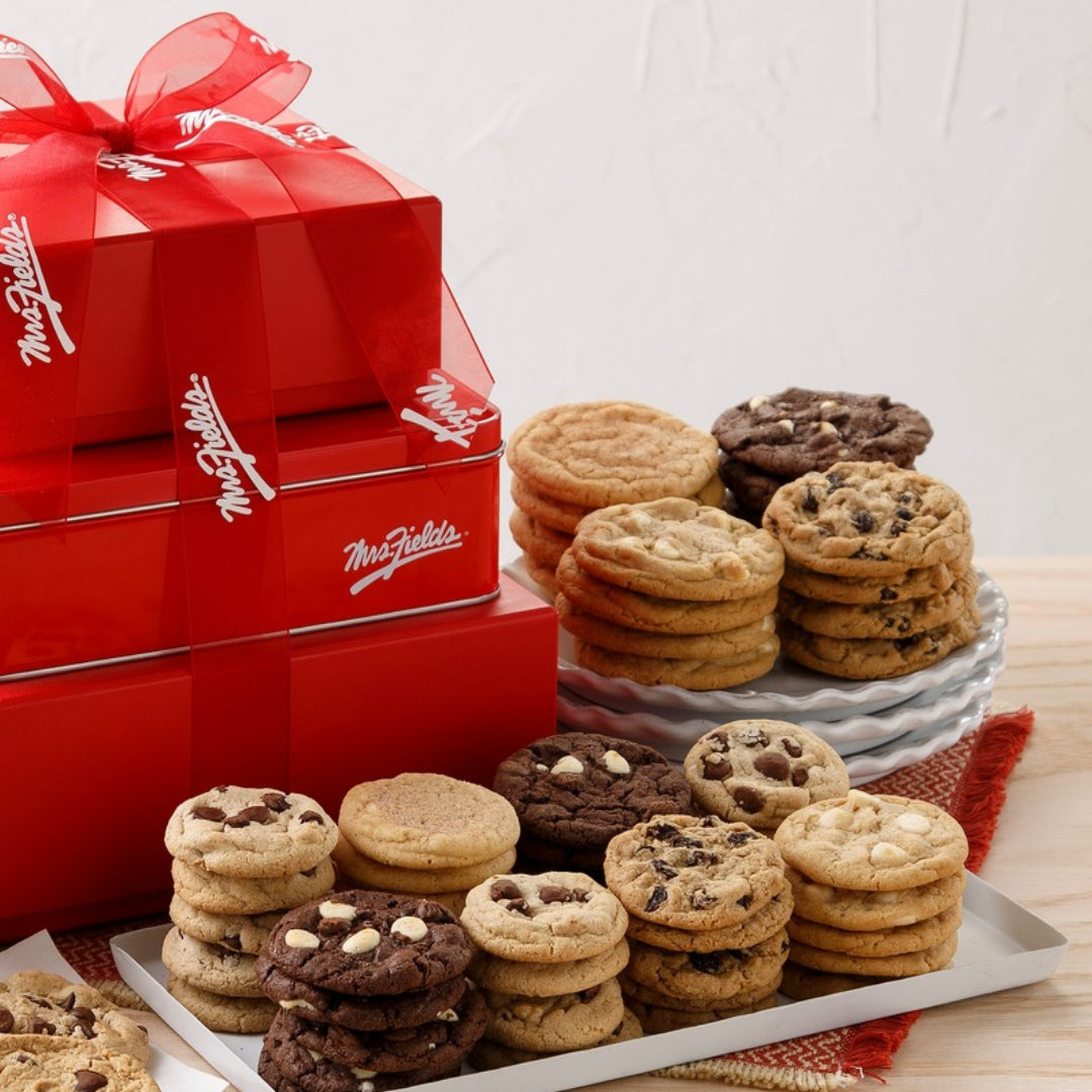 A gift set of chocolate chip cookies and chocolate cookies displayed on white plates and a tray, with three stacked red Mrs. Fields boxes tied with a red ribbon in the background.