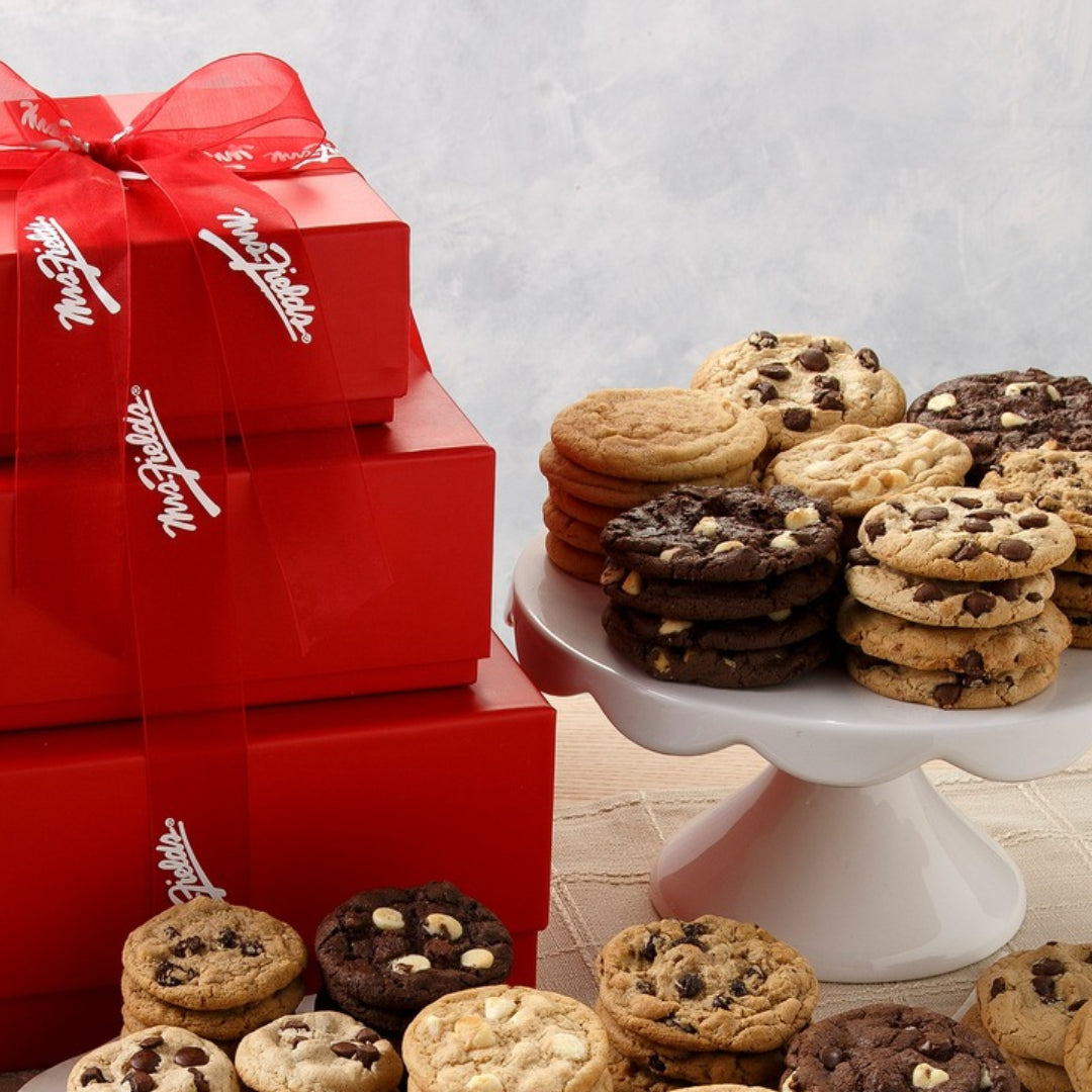 A gift set of chocolate chip cookies and chocolate cookies displayed on white plates and a tray, with three stacked red Mrs. Fields boxes tied with a red ribbon in the background.