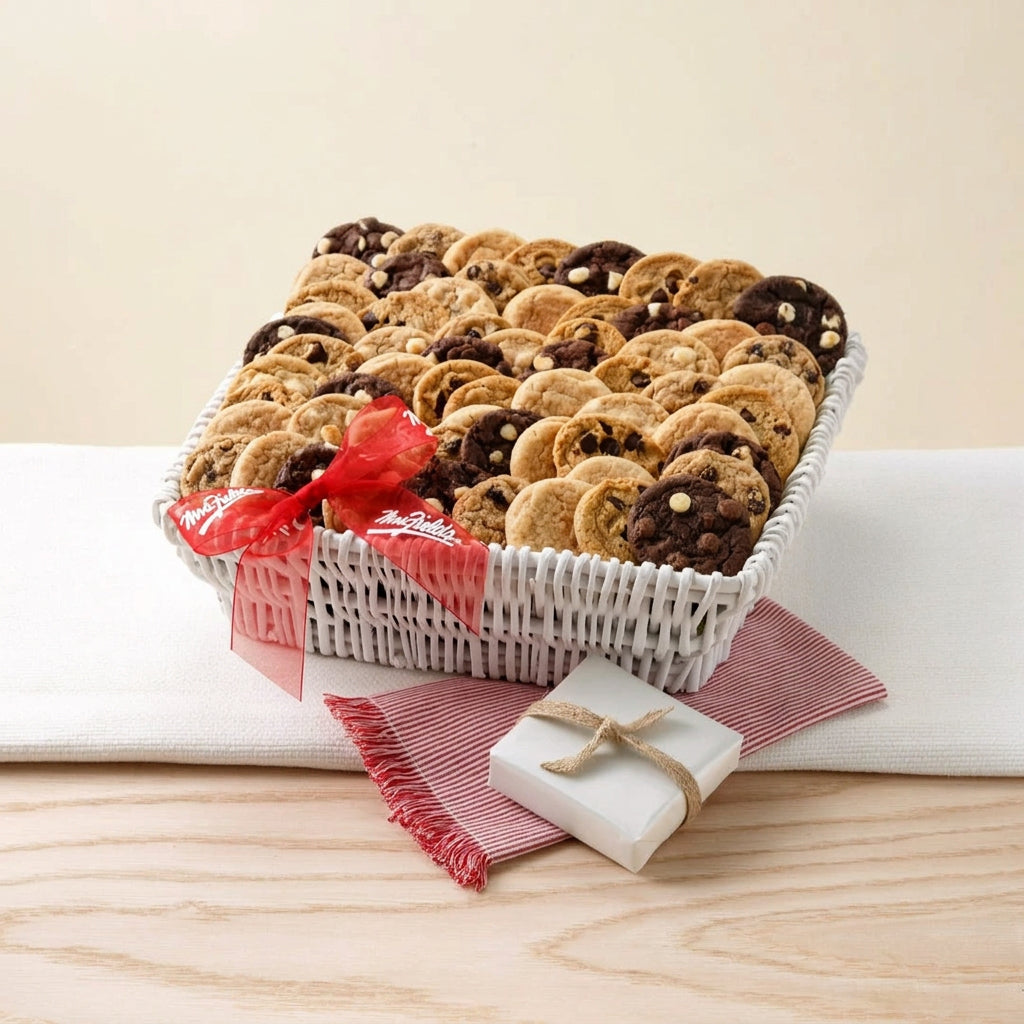 A white woven basket filled with assorted chocolate chip cookies, tied with a red ribbon, displayed on a light wood surface with a white cloth and a small wrapped gift box.