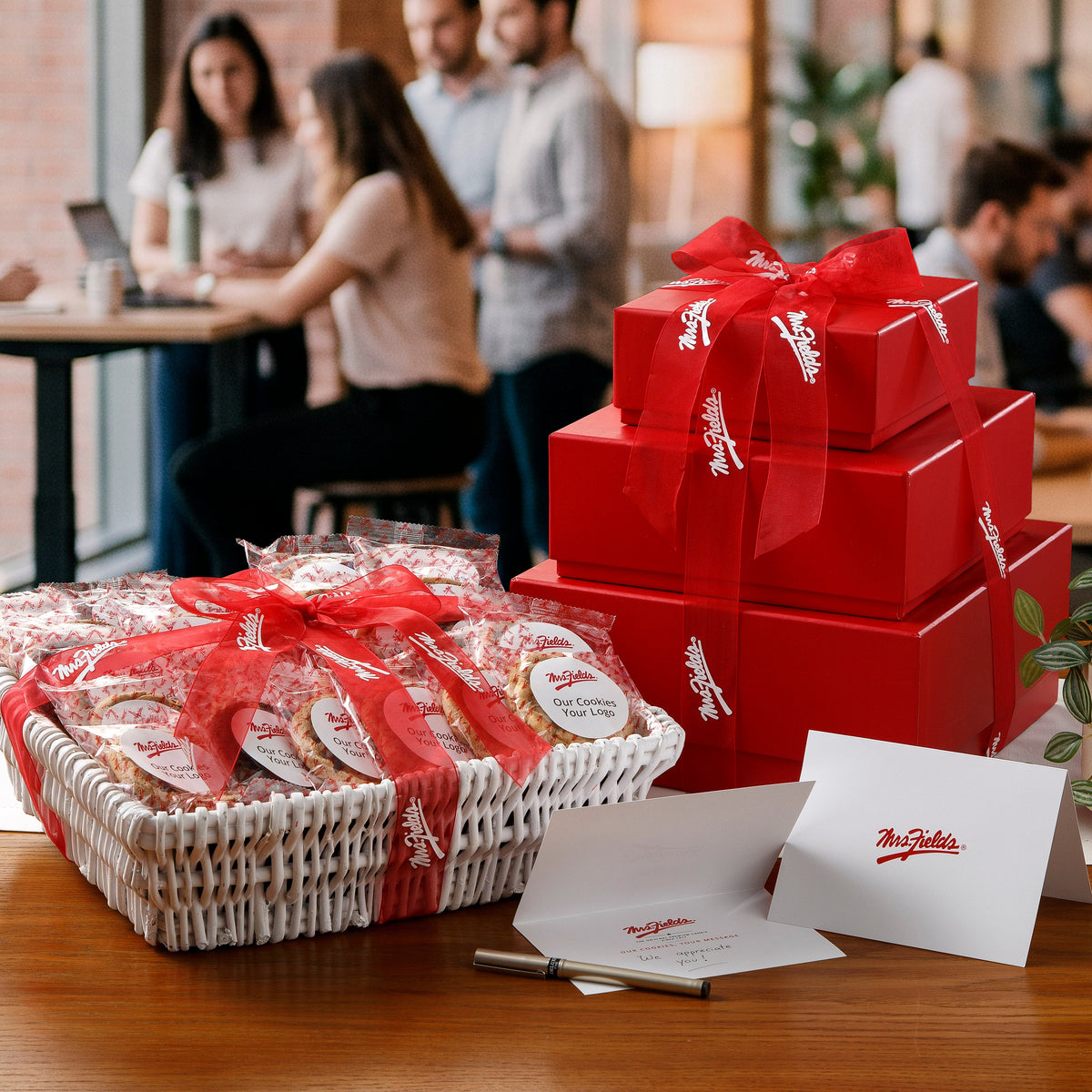 A gift basket filled with packaged cookies tied with a red ribbon sits on a wooden table beside a red gift tower also tied with a red ribbon and Mrs. Fields branded cards, with a blurred office interior in the background.