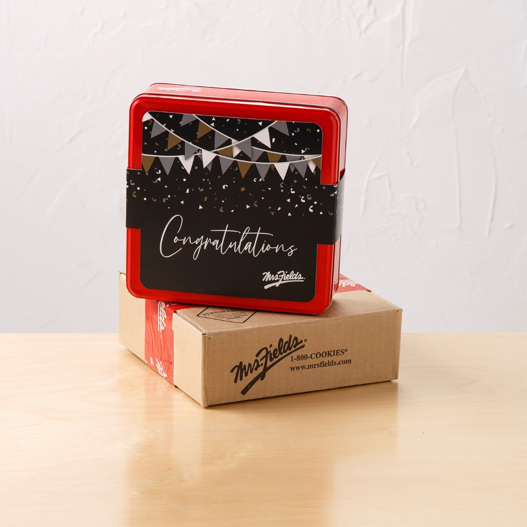 A red square cookie tin with a black 'Congratulations' banner sleeve on top of a brown cardboard mailer box. The tin features a festive print with bunting and confetti, and the brand name 'Mrs. Fields'