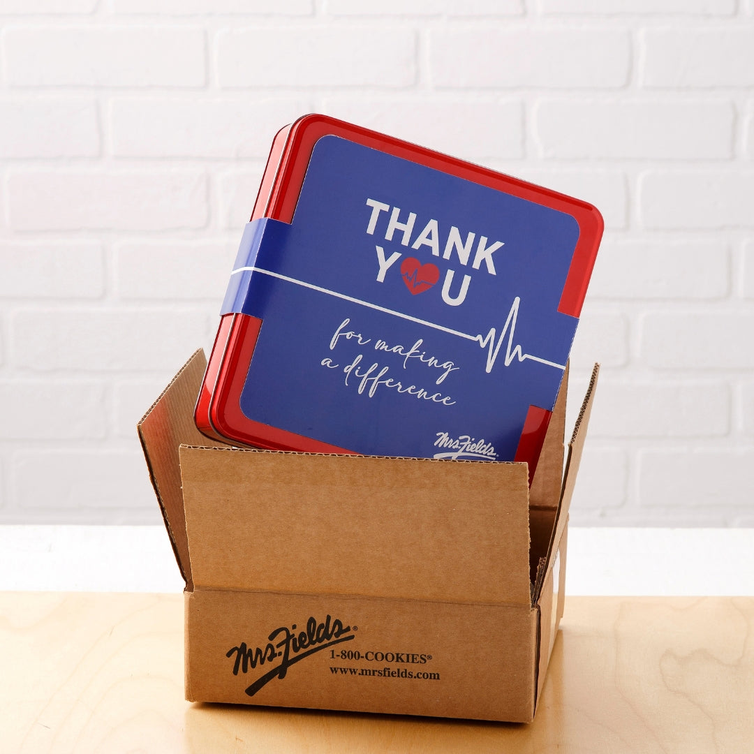 A red and blue square thank-you card or insert displayed inside an open brown cardboard box on a light wood surface, with a white brick wall background. The card reads “THANK YOU for making a difference” and shows the Mrs. Fields logo.