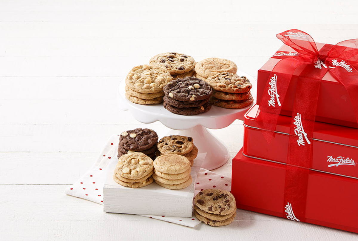 An assortment of chocolate chip cookies and sandwich cookies displayed on white cake stands, next to stacked red gift boxes with white 'The Fight Club' logo and a red ribbon, on a white wood background.