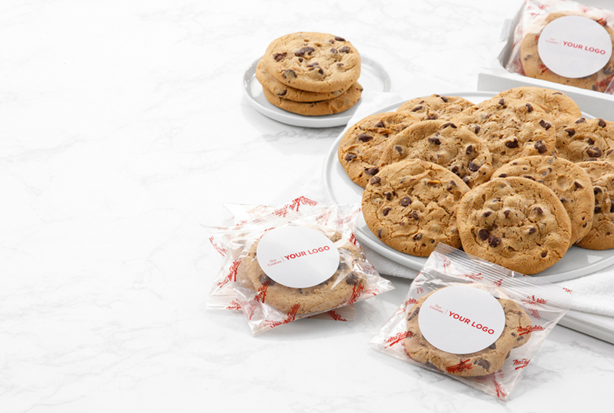 Chocolate chip cookies displayed on white plates and packaged in clear bags with round white labels reading 'YOUR LOGO', arranged on a light marble surface.