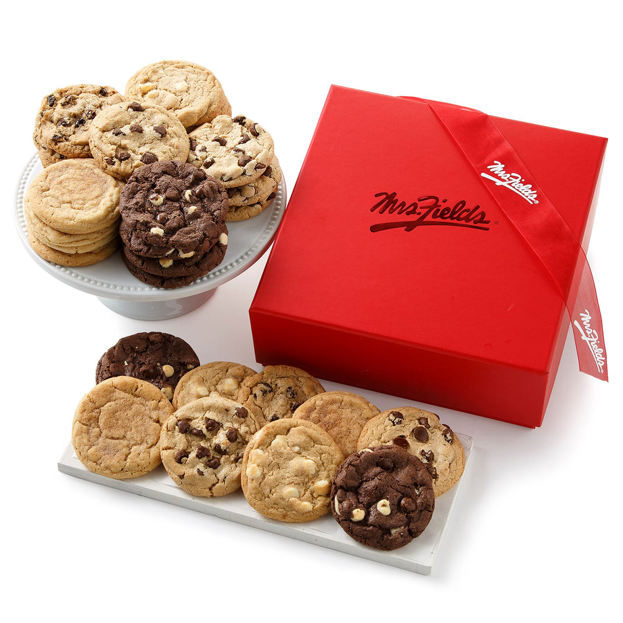 A square red gift box with a shiny Mrs. Fields logo on it surrounded by a large assortment of cookies with a red Mrs. Fields ribbon on a blank white surface.