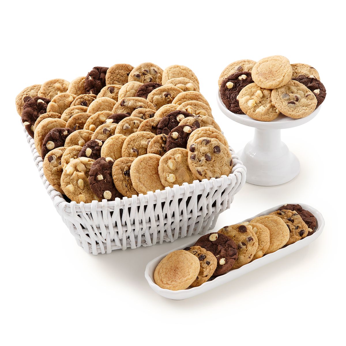 A large assortment of chocolate chip cookies in a white woven basket, with additional cookies displayed on a white pedestal stand and a white oval platter against a clean white background.