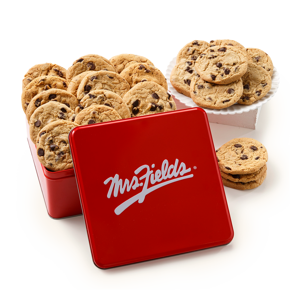 A red square metal cookie tin with the white MrsFields logo on the lid, shown with chocolate chip cookies inside and a few cookies stacked on a white plate beside it on a clean white background.