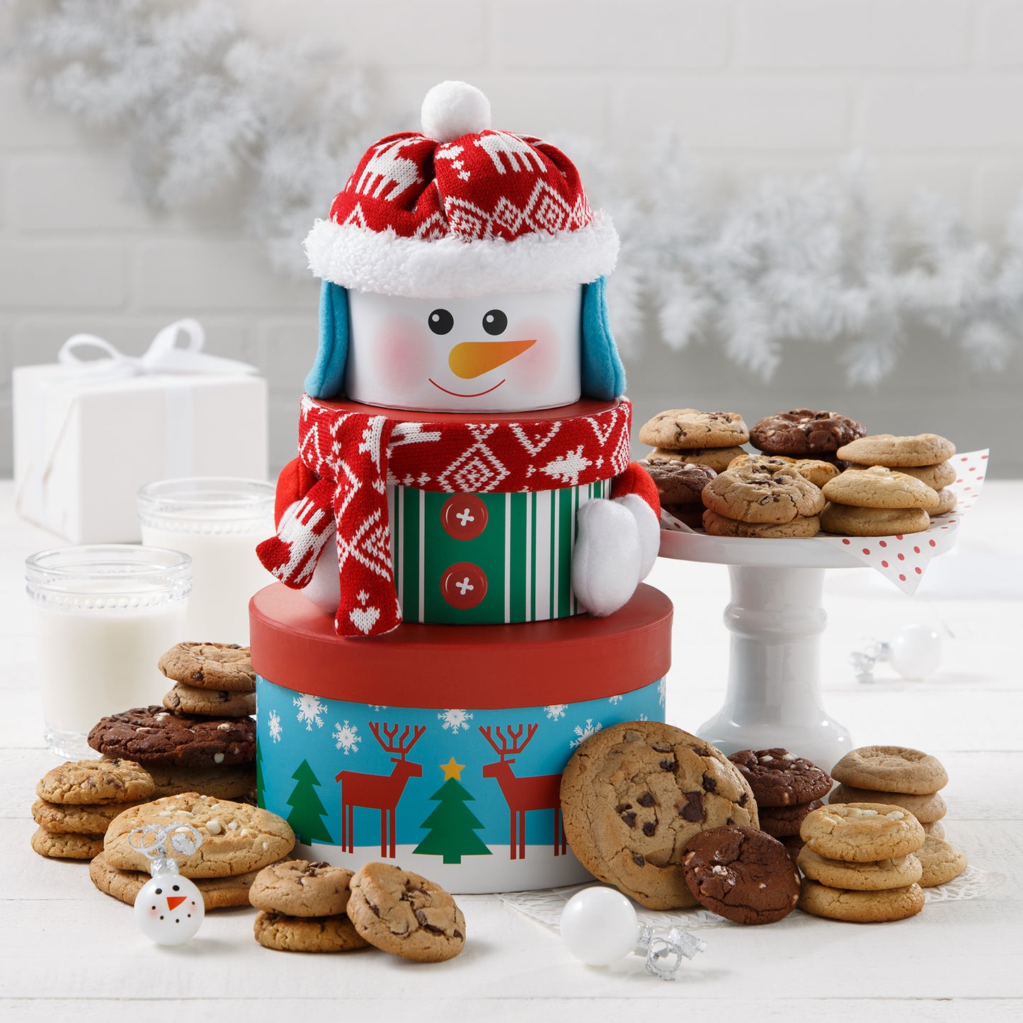 A cute three-tiered tower of round boxes that make a snowman when stacked together wearing a knitted cap and scarf and surrounded by an assortment of Mrs. Fields cookies in five flavors including chocolate chip and white chocolate macadamia nut on a white surface with two glasses of milk, a white garland, and white Christmas ornaments.