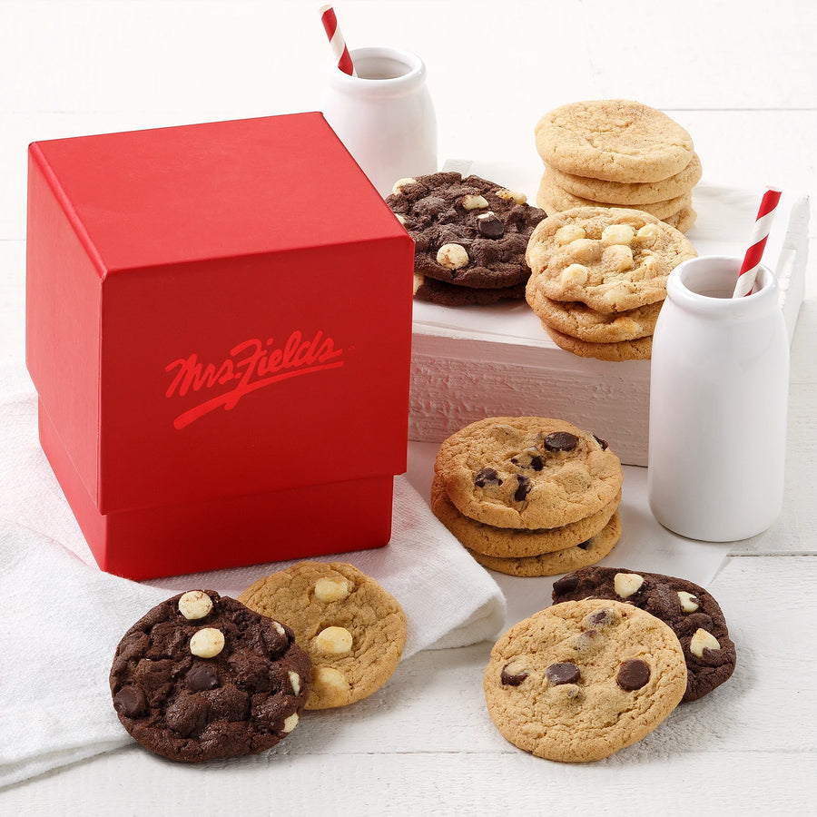 A red gift box with a shiny Mrs. Fields logo on it surrounded by an assortment of Nibblers bite-sized cookies on a white wooden surface with two milk bottles that have white and red striped straws in them.