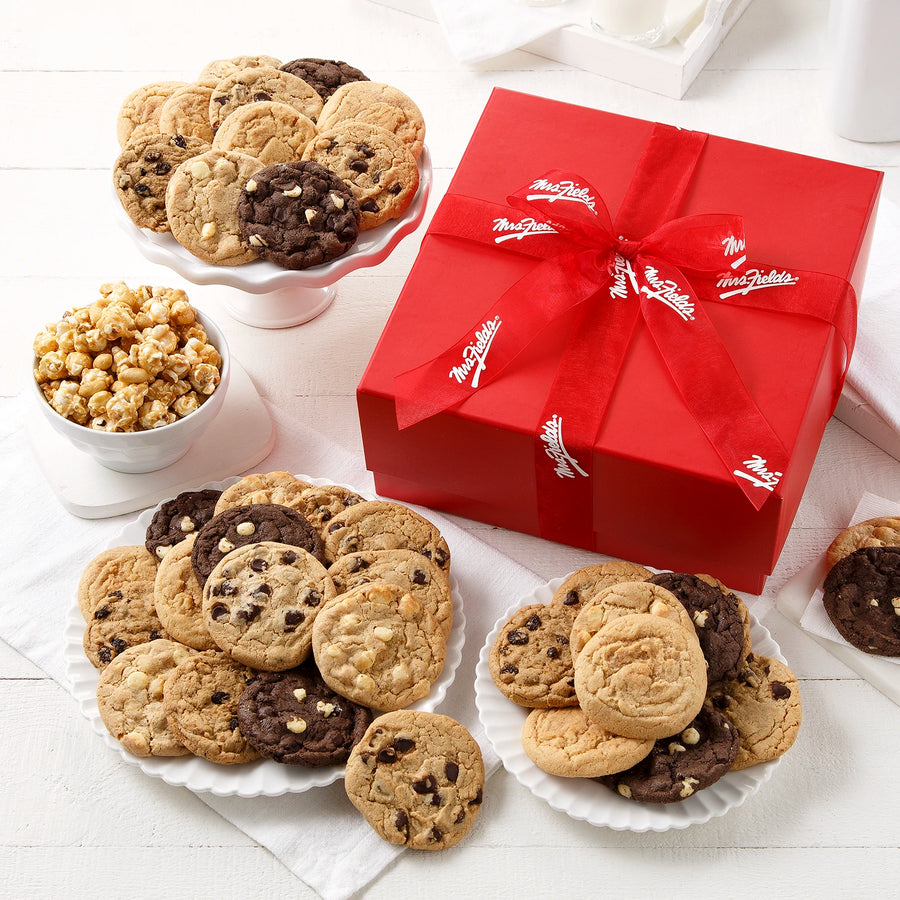 Red gift box tied with a Mrs. Fields red ribbon surrounded by an assortment of 38 Mrs. Fields cookies and toffee nut popcorn on a white surface with white dishes.