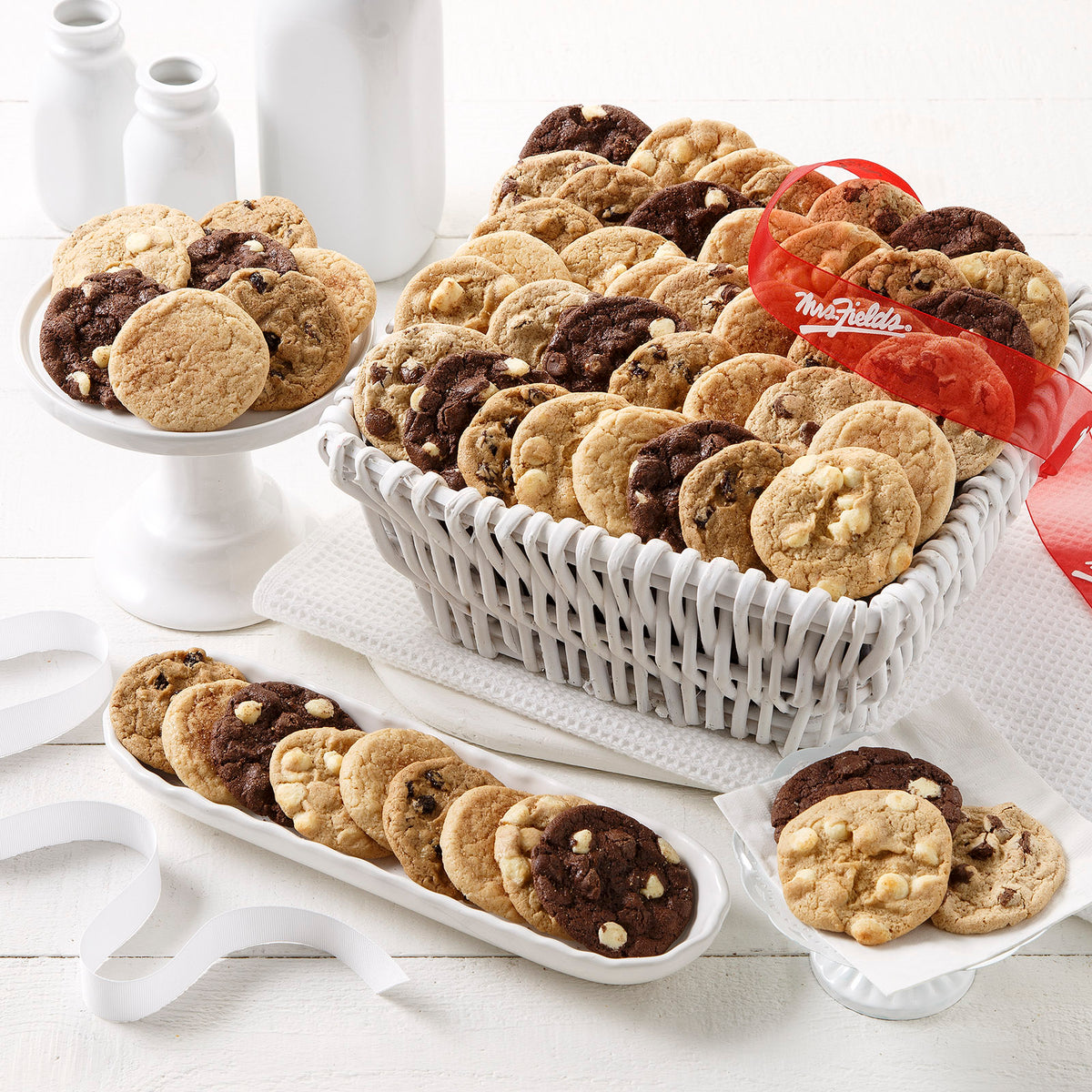 A white wicker basket filled with assorted cookies, including chocolate chip and oatmeal cookies, displayed on a white table with white ceramic dishes and bottles. A red ribbon with white script is tied around the basket.