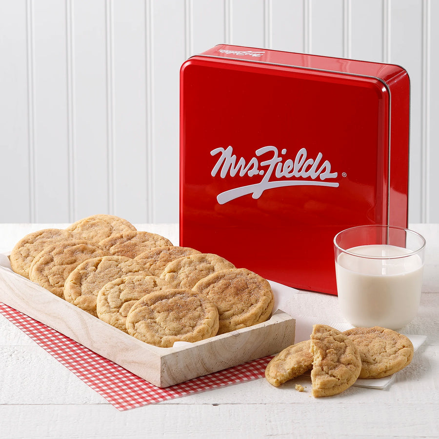 Mrs. Fields cinnamon sugar cookies in a wooden tray and scattered on a table, with a glass of milk and a red cookie tin featuring the white Mrs. Fields logo