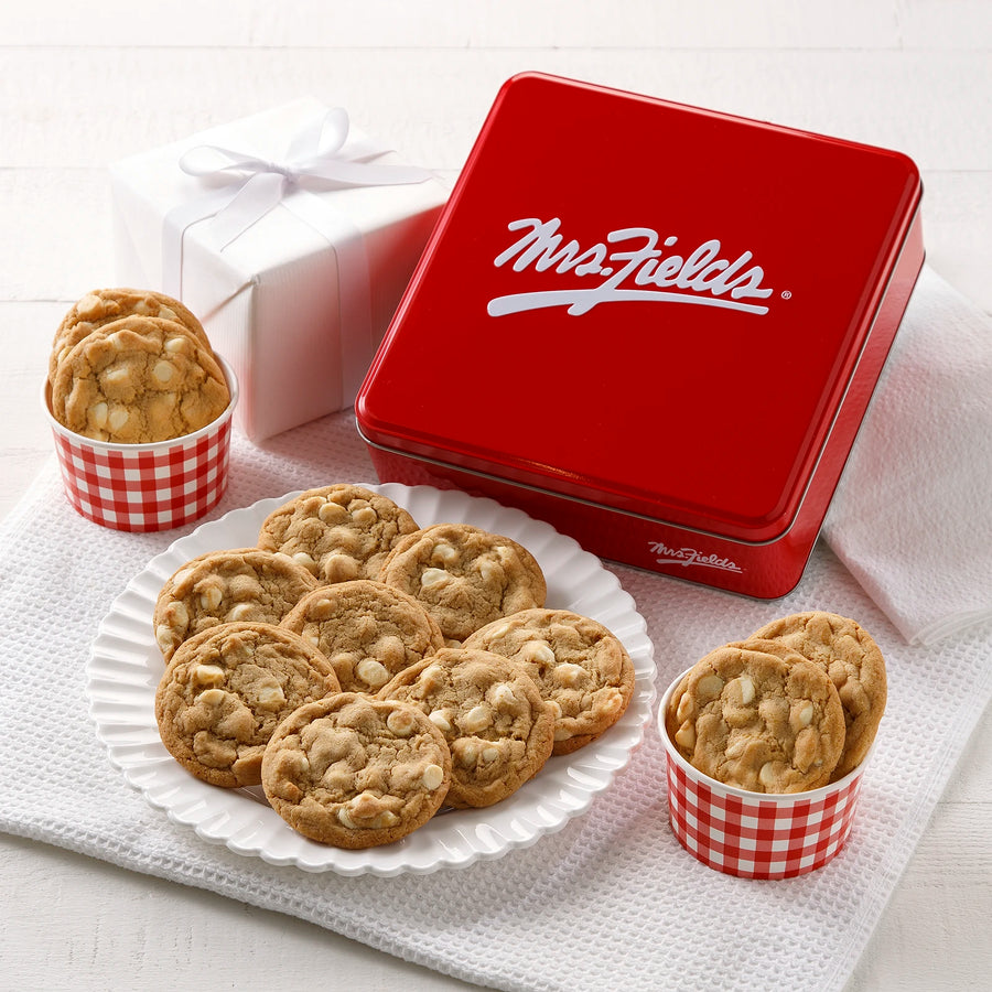 A red Mrs. Fields cookie tin with a dozen white chocolate chip cookies on a white plate and in two red gingham bowls, all on a white cloth with a white gift wrapped next to it.