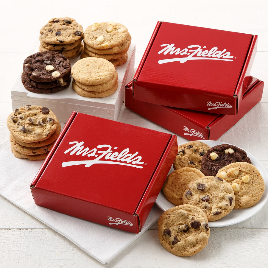 Three red boxes with a white Mrs. Fields logo on them stacked and surrounded by an assortment of small cookies on a white wooden surface.