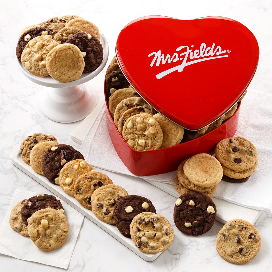 A heart shaped red Mrs. Fields cookie tin filled with an assortment of cookies all on a white surface with white napkins and plates.