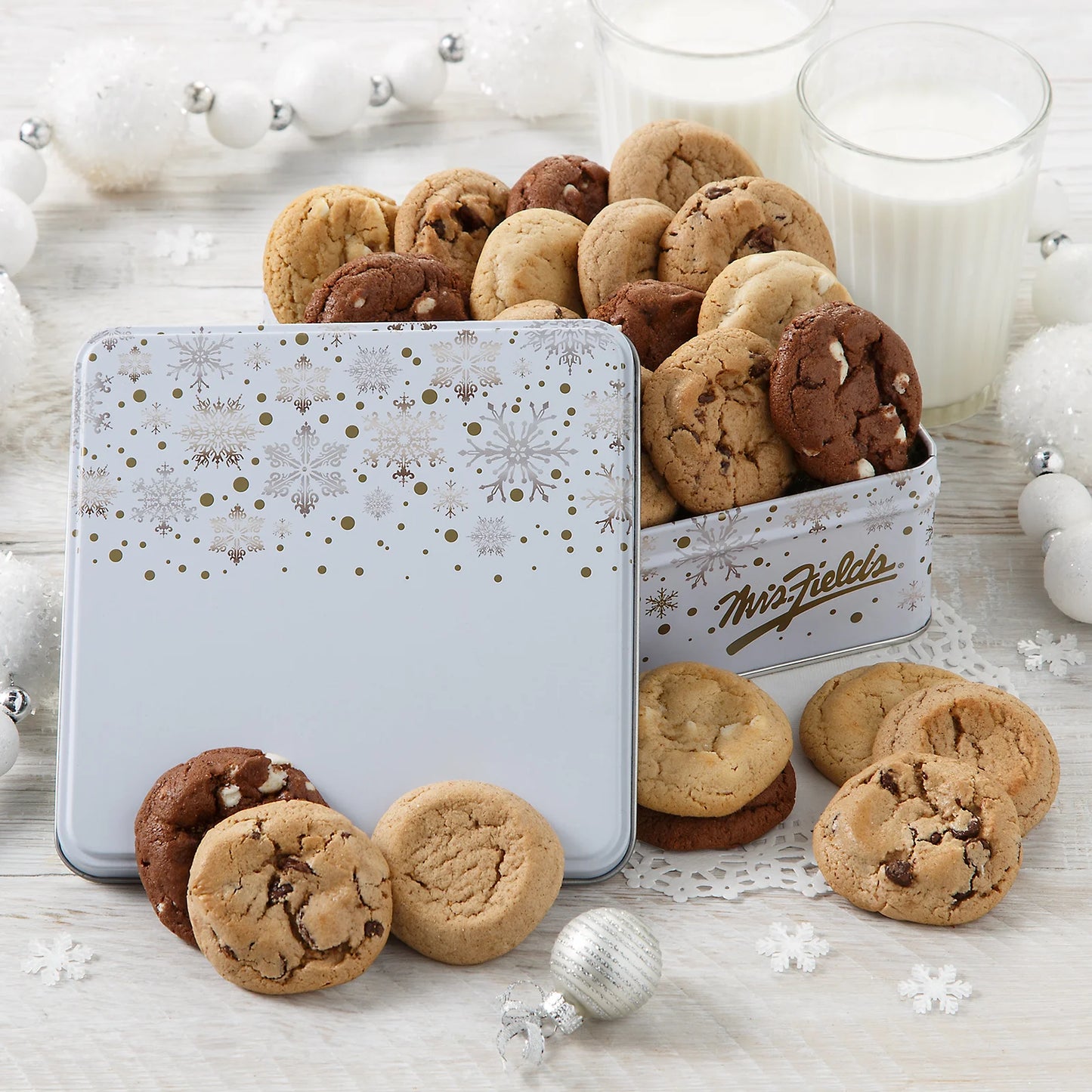 Assorted Mrs. Fields Nibblers bite-sized cookies in a holiday themed tin with a decorative snowflake lid on a light wooden surface with a silver garland and two glasses of milk.