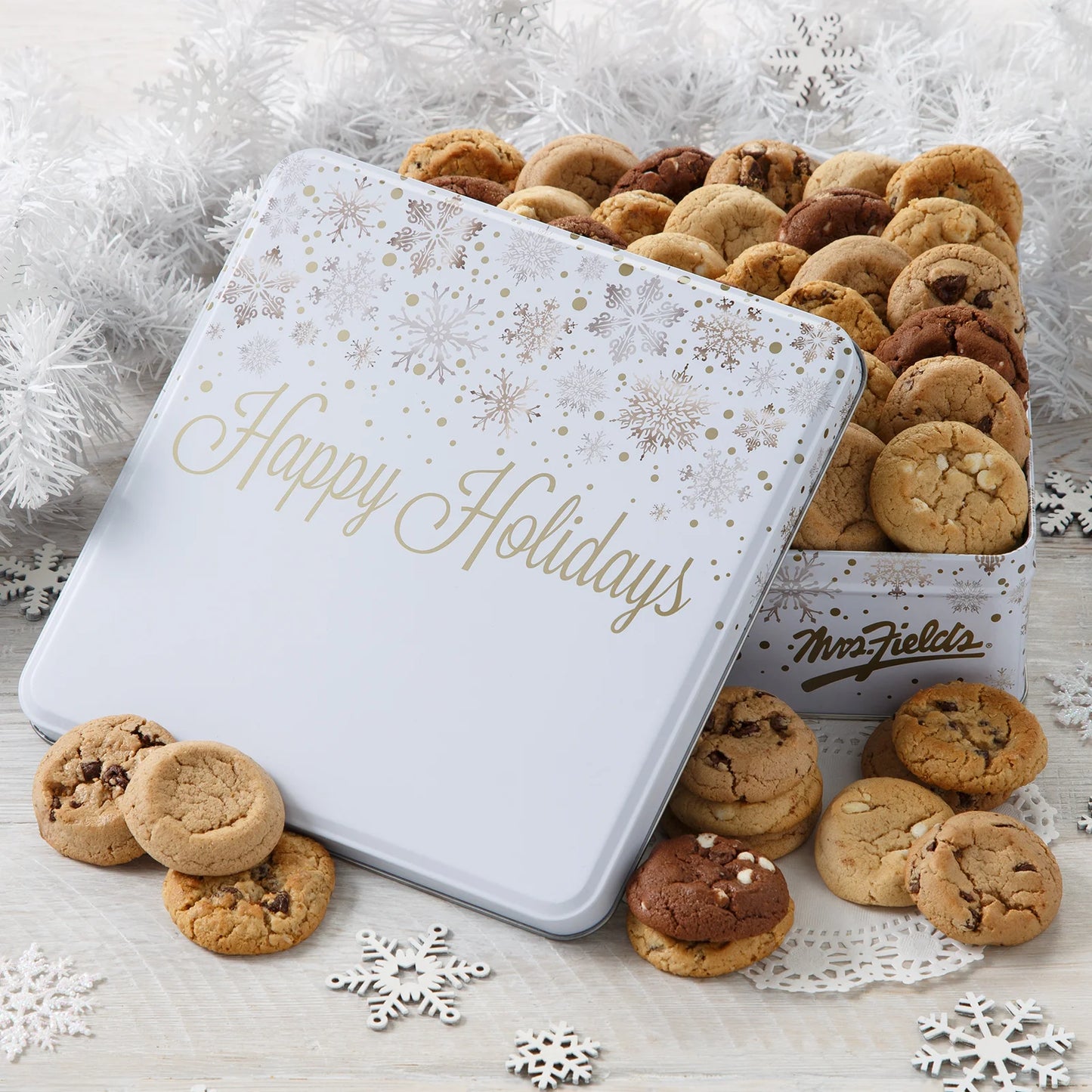 A "Happy Holidays" snowflake pattern cookie tin filled with Mrs. Fields Nibblers bite-sized cookies in five flavors including chocolate chip and triple chocolate on a white surface with snowflake cutouts