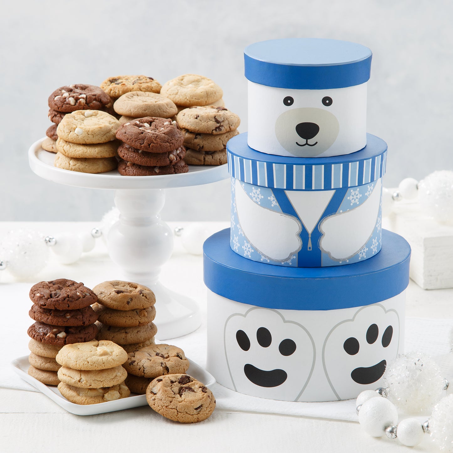 A three-tiered blue and white tower of round boxes that make a polar bear when stacked together surrounded by an assortment of Mrs. Fields cookies in five flavors including chocolate chip and cinnamon sugar on a white surface with white Christmas ornaments.