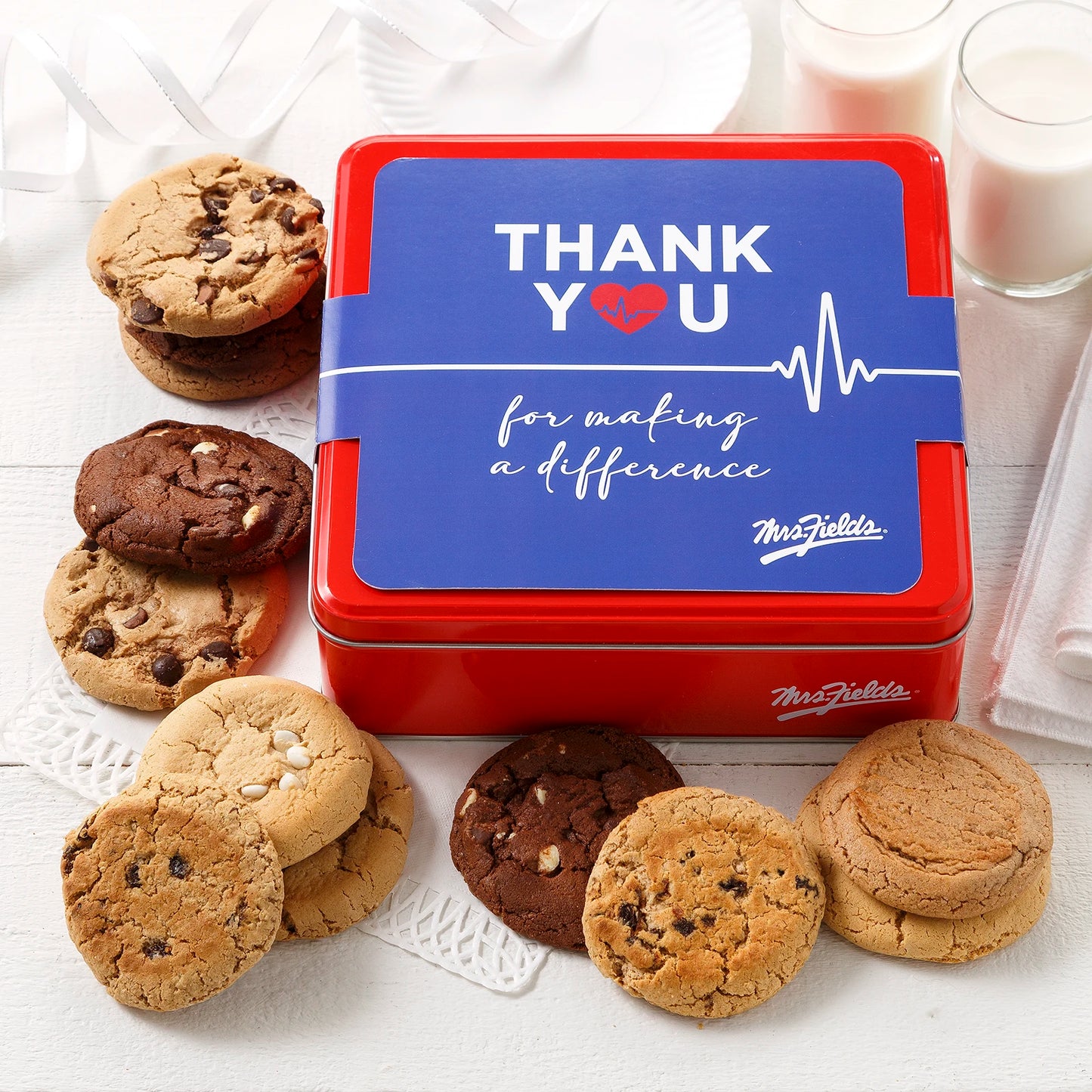 Mrs. Fields signature red cookie tin with a "Thank You for making a difference" sleeve on it surrounded by a dozen original cookies in five flavors including chocolate chip and white chocolate chip on a white surface with two glasses of milk.