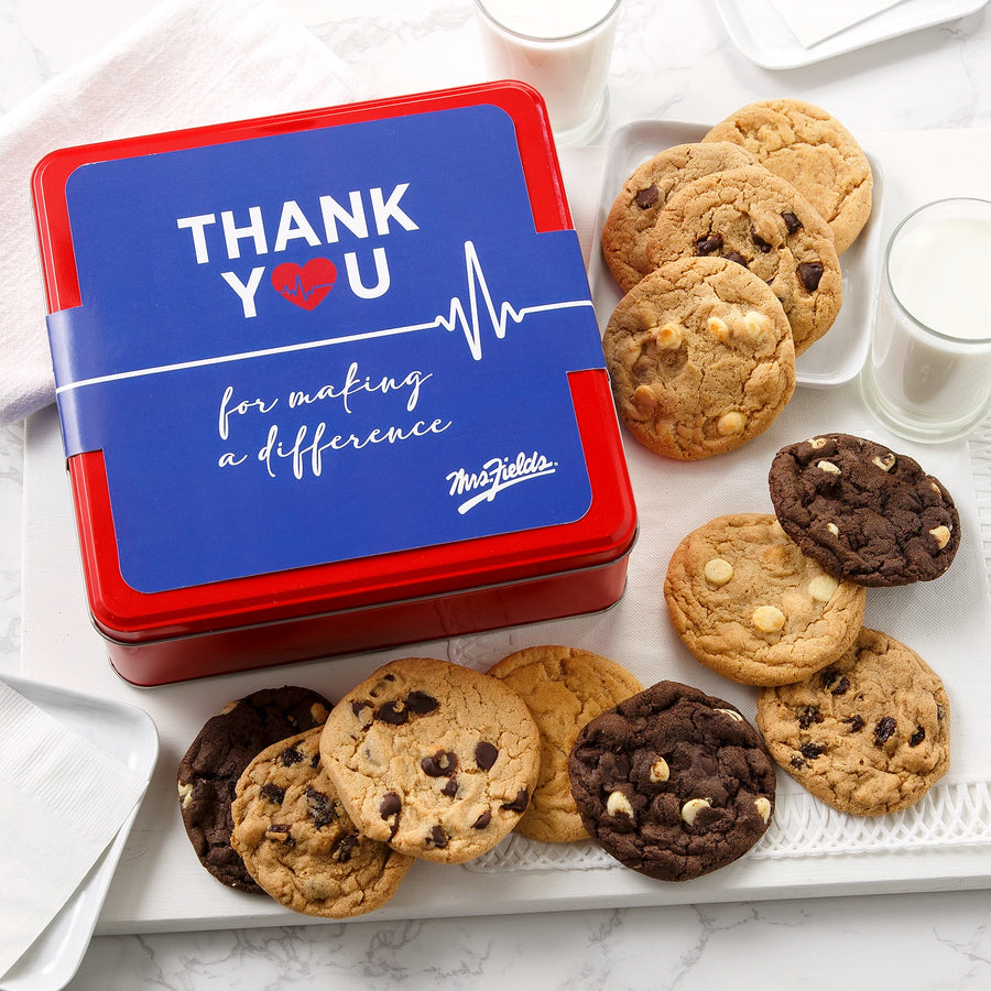 A red Mrs. Fields cookie tin with a blue sleeve on it that says "Thank You for making a difference" with a heart beat graphic on it surrounded by an assortment of a dozen cookies and two glasses of milk on a white surface.