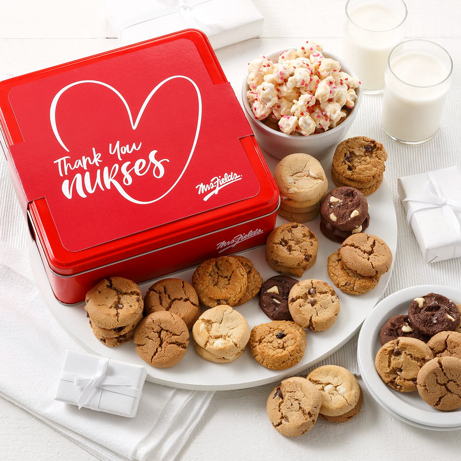 Mrs. Fields signature red cookie tin with a white sleeve that says "Thank You Nurse" on it surrounded by an assortment of Nibblers bite-sized cookies and popcorn with heart sprinkles on a white surface with two glasses of milk.