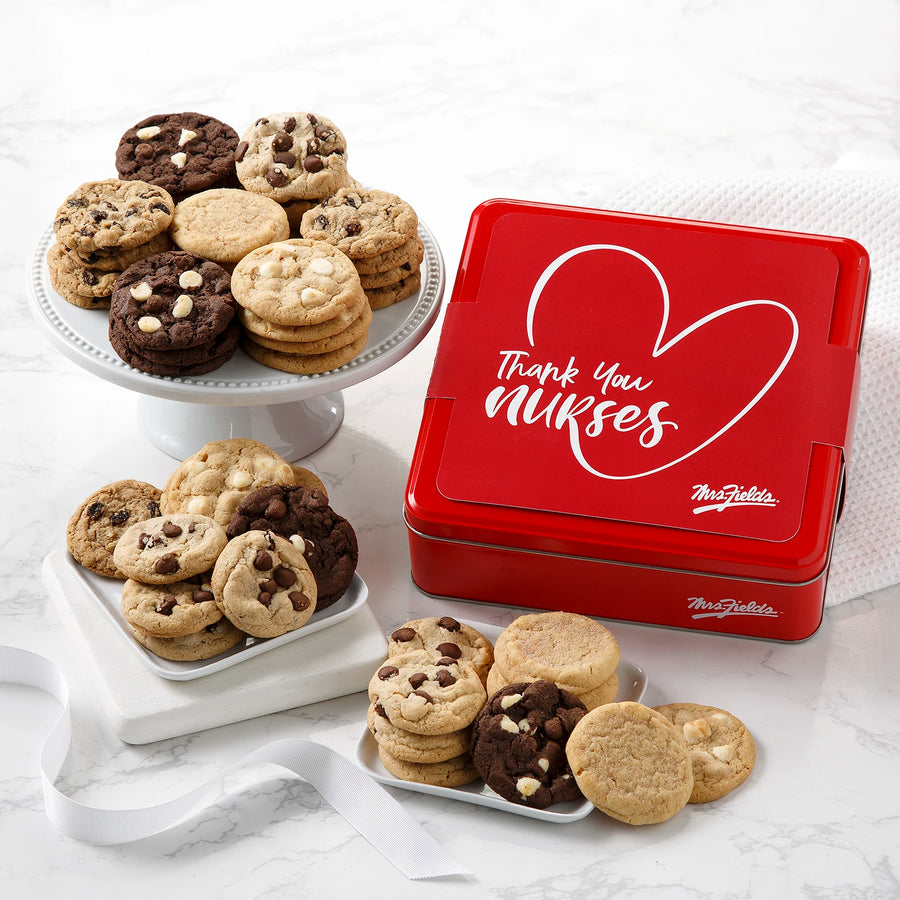 A red Mrs. Fields cookie tin with a red sleeve on it that says "Thank you nurses" with a heart graphic surrounded by an assortment of Nibblers bite-sized cookies on a white marbled surface with white plates and a white ribbon.