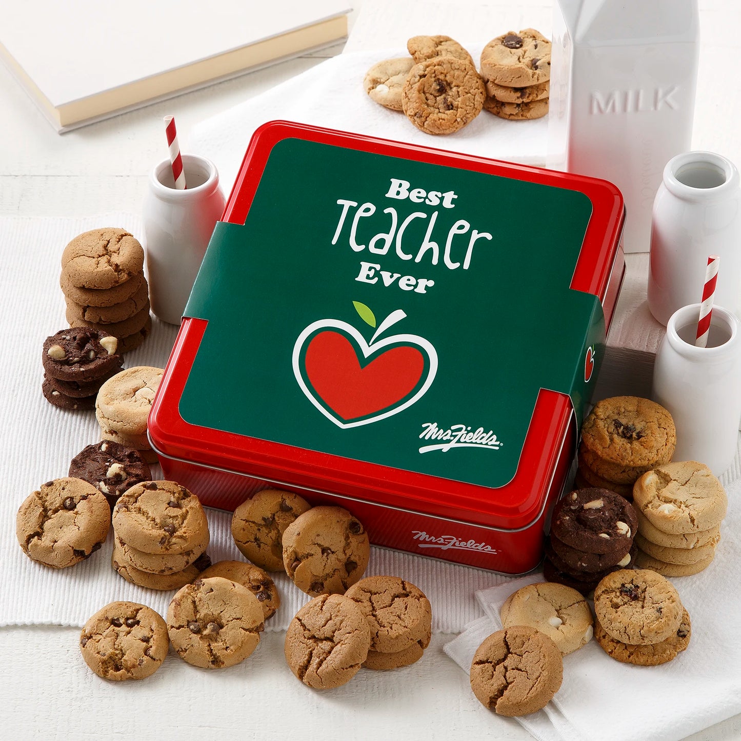 A Mrs. Fields signature red tin with a "Best Teacher Ever" sleeve on it surrounded by an assortment of Nibblers bite-sized cookies on a white surface.