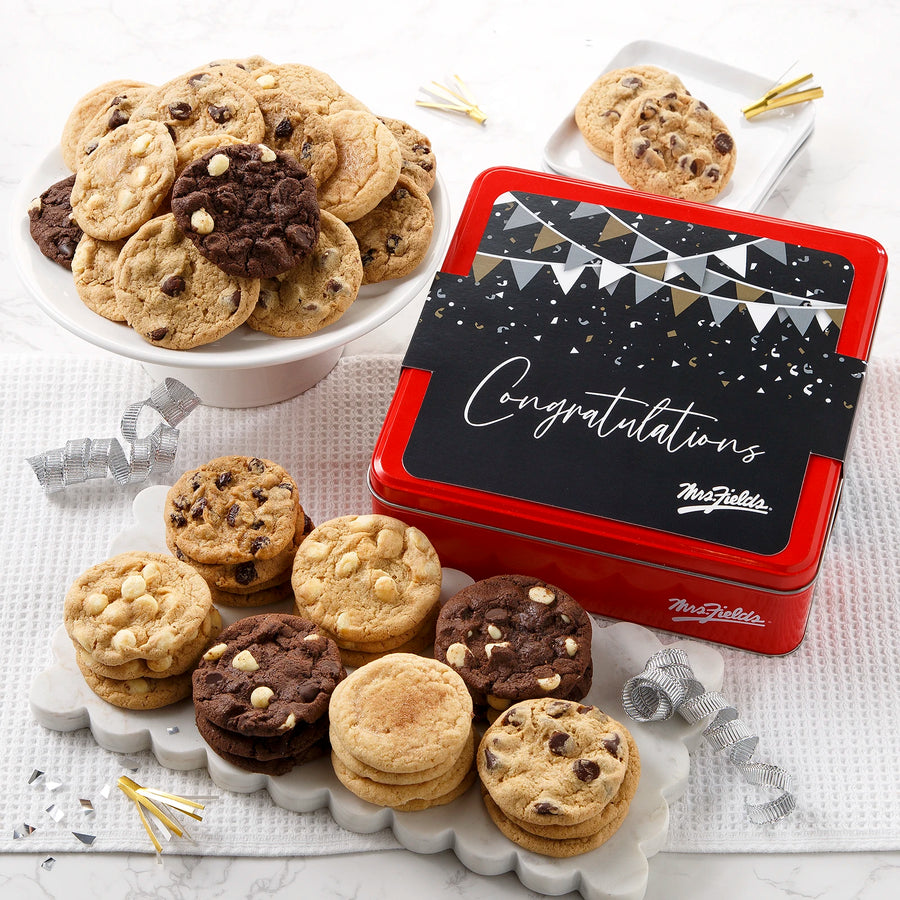 A Mrs. Fields red cookie tin with a black, silver, and gold sleeve on it that says "Congratulations" in a cursive font surrounded by an assortment of Nibblers bite-sized cookies on a white surface with ribbon and confetti.