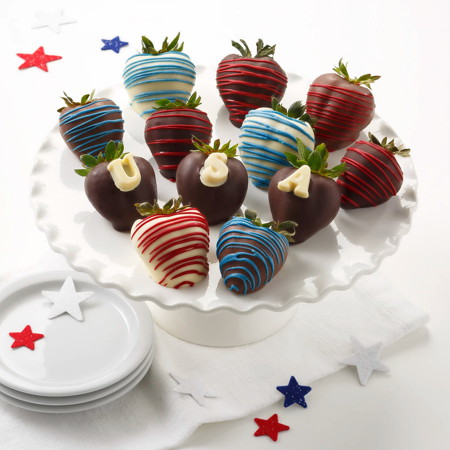 Twelve chocolate covered strawberries decorated in red, white, and blue chocolate drizzles and white chocolate lettering spelling "USA" on a white surface with star decorations.
