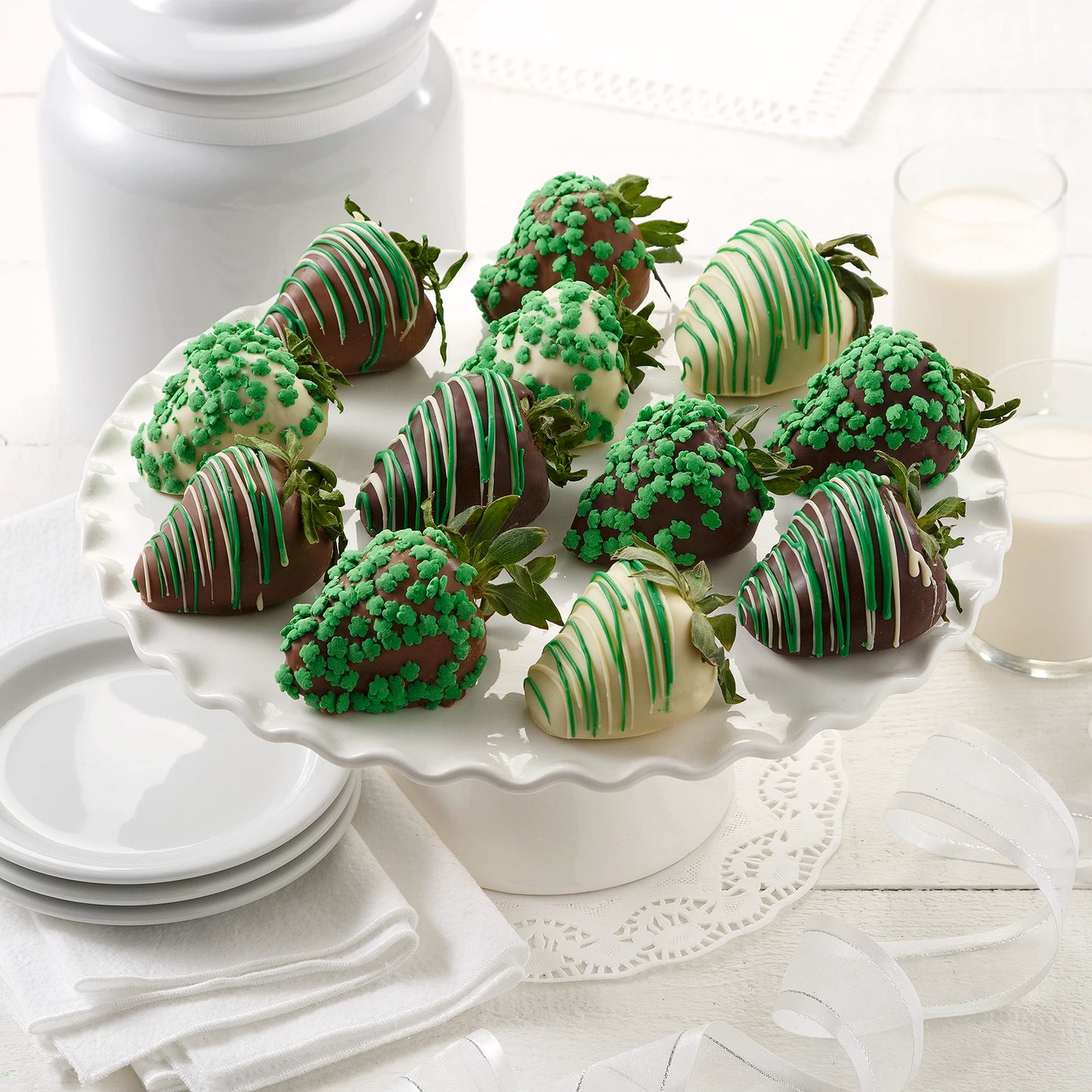 Twelve St. Patrick's Day themed chocolate covered strawberries with green chocolate drizzles and shamrock sprinkles on a white platter with white dishes and glasses of milk.