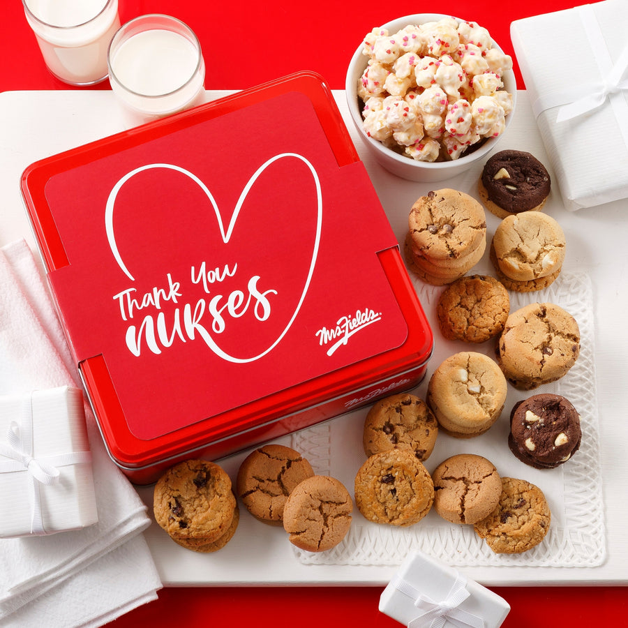 Mrs. Fields signature red cookie tin with a white sleeve that says "Thank You Nurses" on it surrounded by an assortment of Nibblers bite-sized cookies and popcorn with heart sprinkles on a white tray on a red surface with two glasses of milk, napkins, and small wrapped gifts.