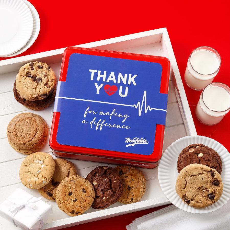 Mrs. Fields signature red cookie tin with a "Thank You for making a difference" sleeve on it surrounded by a dozen original cookies in a white tray on a red surface with plates and two glasses of milk.