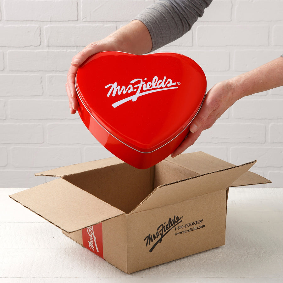 Mrs. Fields signature heart shaped tin coming out of a Mrs. Fields branded mailer on a white table top surface.
