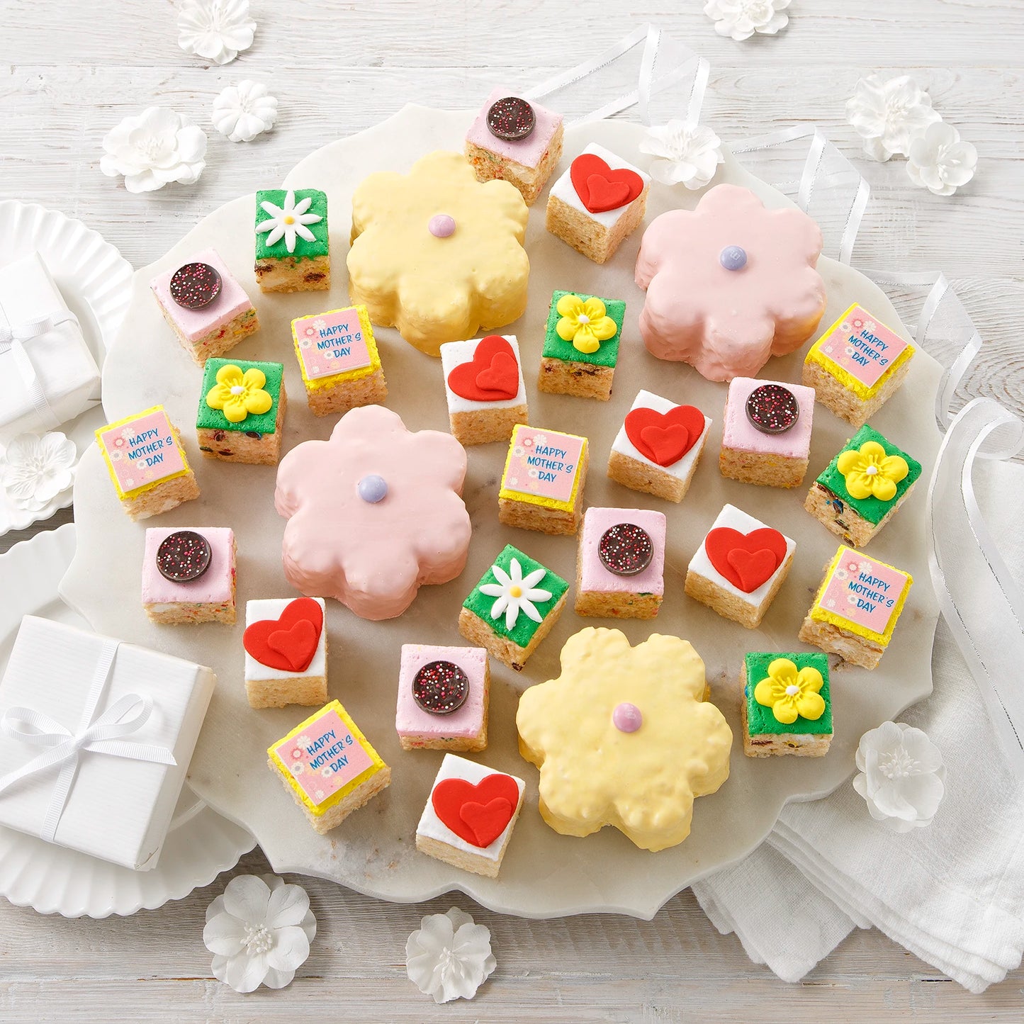 An assortment of rice krispie treats including four large flower shaped treats and two dozen mini treats all decorated with a springtime and Mother's Day theme on a gray surface with white dishes.