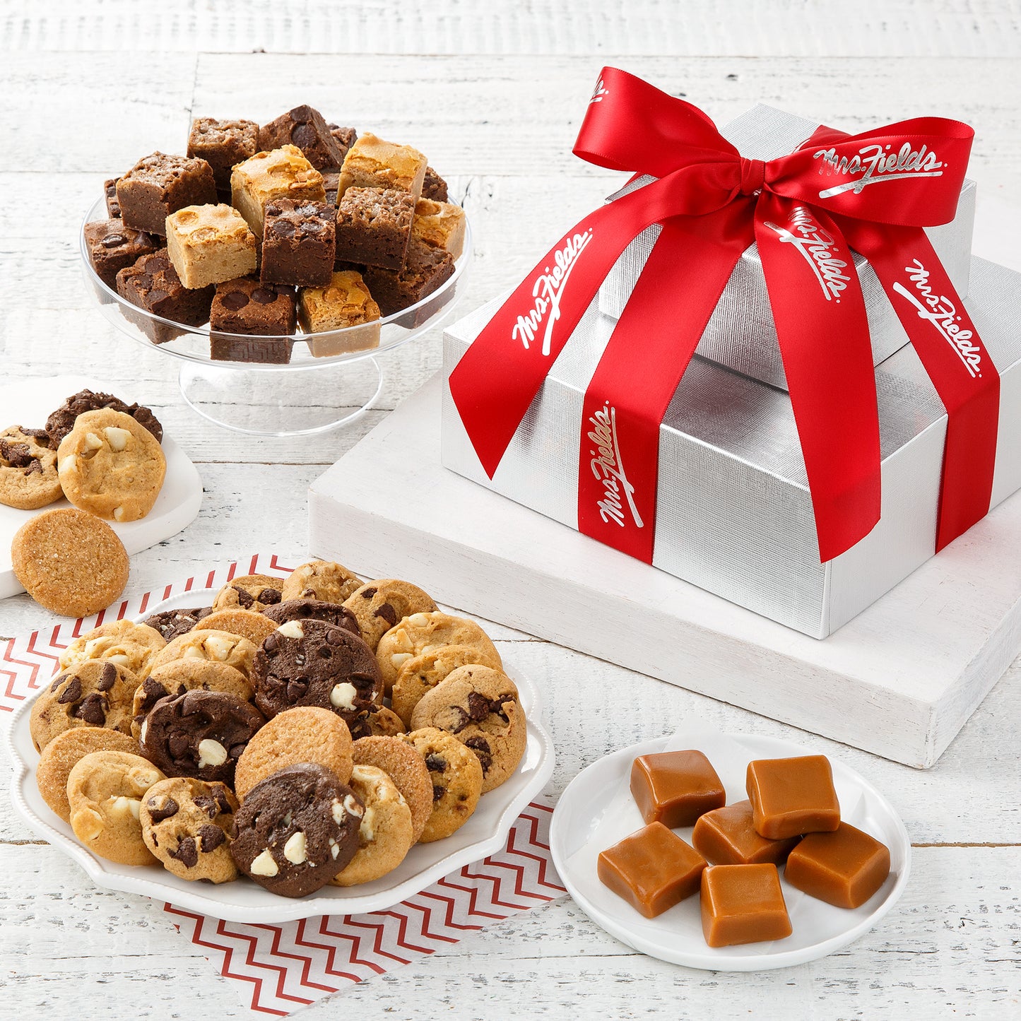 Two tier silver box tower tied with a red Mrs. Fields ribbon and surrounded by an assortment of Nibblers® and brownie bites.