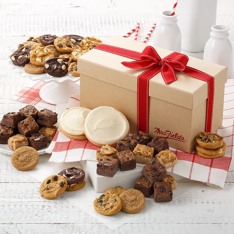 A brownie gift box tied with a red ribbon surrounded by an assortment of nibblers, brownies bites, and frosted cookies