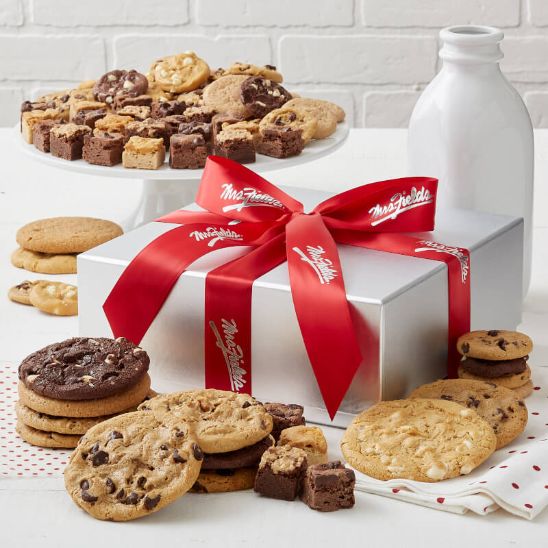 A silver gift box and tied with a red Mrs. Fields ribbon and surrounded by an assortment of Nibblers®, original cookies, and brownie bites
