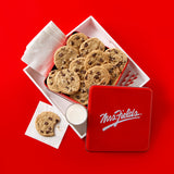 A red tin of Mrs. Fields chocolate chip cookies arranged in an open white box on a bright red background, with a few cookies on napkins and a small cup of milk beside the box.