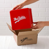 A person's hands are shown lifting a square, glossy red metal tin with a white Mrs. Fields logo out of brown cardboard box branded with Mrs. Fields and 1-800-COOKIES. The background is a clean white brick wall.